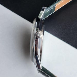 May include: A silver wristwatch with a green and tan leather strap. The watch face is round and the side view shows a textured crown. The watch is set against a black and white background.