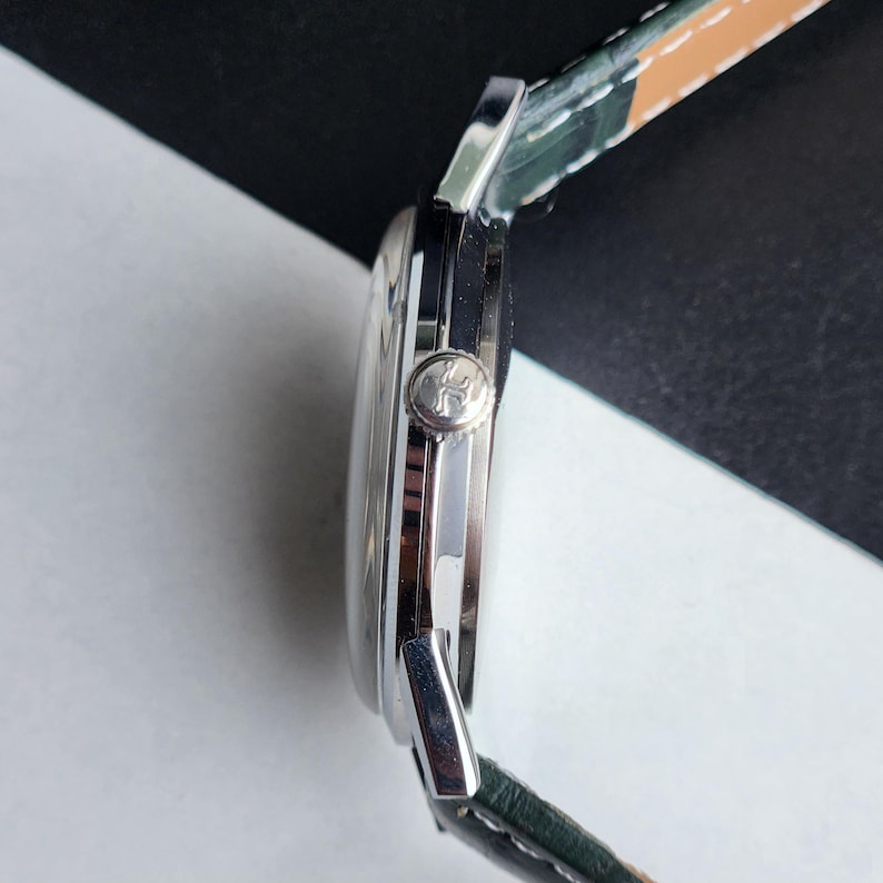 May include: A silver wristwatch with a green and tan leather strap. The watch face is round and the side view shows a textured crown. The watch is set against a black and white background.