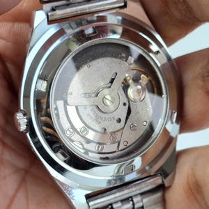 K&ouml;nnte beinhalten: Nahaufnahme einer silbernen Armbanduhr mit transparentem Geh&auml;useboden, der das komplizierte mechanische Uhrwerk zeigt. Die Uhr hat ein silbernes Armband und ein rundes Zifferblatt. Die Worte "JAPAN SEVENTEEN" sind auf dem Uhrwerk zu sehen.
