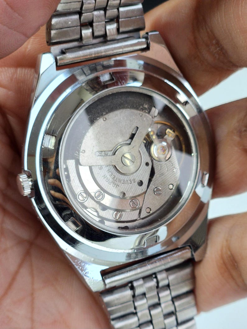 K&ouml;nnte beinhalten: Nahaufnahme einer silbernen Armbanduhr mit transparentem Geh&auml;useboden, der das komplizierte mechanische Uhrwerk zeigt. Die Uhr hat ein silbernes Armband und ein rundes Zifferblatt. Die Worte "JAPAN SEVENTEEN" sind auf dem Uhrwerk zu sehen.