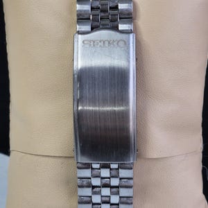 K&ouml;nnte beinhalten: Nahaufnahme eines Edelstahl-Uhrenarmbands mit geb&uuml;rsteter Metallschnalle. Auf der Schnalle ist der Markenname "SEIKO" eingraviert. Das Armband hat ein gitterartiges Muster aus rechteckigen Gliedern.