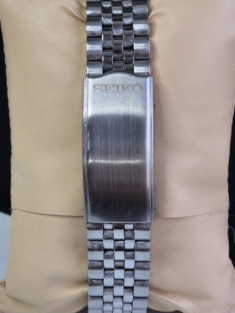 K&ouml;nnte beinhalten: Nahaufnahme eines Edelstahl-Uhrenarmbands mit geb&uuml;rsteter Metallschnalle. Auf der Schnalle ist der Markenname "SEIKO" eingraviert. Das Armband hat ein gitterartiges Muster aus rechteckigen Gliedern.