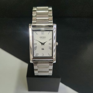 May include: A rectangular Seiko Quartz wristwatch with a silver metal strap and case. The white watch face has black Roman numerals, black hands, and the Seiko Quartz logo. The watch is displayed on a black surface.