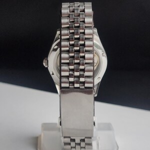 Puede incluir: Un reloj de pulsera plateado con una correa de acero inoxidable. La esfera del reloj es visible a trav&eacute;s de la parte trasera transparente. La correa est&aacute; compuesta por peque&ntilde;os eslabones rectangulares. El reloj se muestra sobre un fondo blanco y negro.