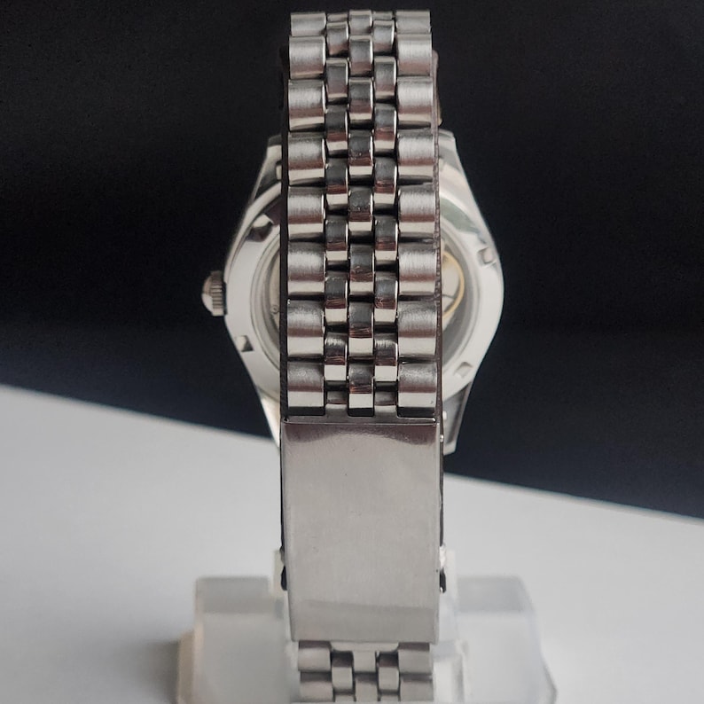 Puede incluir: Un reloj de pulsera plateado con una correa de acero inoxidable. La esfera del reloj es visible a trav&eacute;s de la parte trasera transparente. La correa est&aacute; compuesta por peque&ntilde;os eslabones rectangulares. El reloj se muestra sobre un fondo blanco y negro.