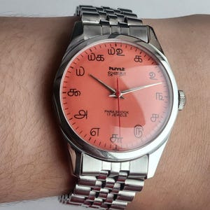 Puede incluir: Un reloj de pulsera plateado con esfera color salm&oacute;n. El reloj tiene una correa de metal y presenta texto en un idioma desconocido. La esfera del reloj incluye el texto "PARA SHOCK 17 JEWELS."