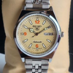 Könnte beinhalten: Eine silberfarbene Seiko 5 Automatik-Armbanduhr mit hellgoldenem Zifferblatt. Die Uhr verfügt über eine Tages- und Datumsanzeige, Leuchtzeiger und ein Edelstahlarmband. Die Ziffer 12 ist rot.