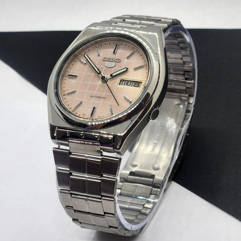 May include: A silver-toned Seiko 5 automatic wristwatch with a pink, grid-patterned face. The watch features a day-date display and a stainless steel band. The watch face displays the day as "THU 8".