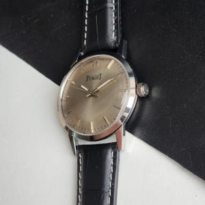 Puede incluir: Un reloj Piaget plateado con esfera marr&oacute;n claro y correa de cuero negro. El reloj tiene una esfera redonda con agujas e indicadores plateados. La correa es de cuero negro con costuras blancas. Dise&ntilde;o cl&aacute;sico.