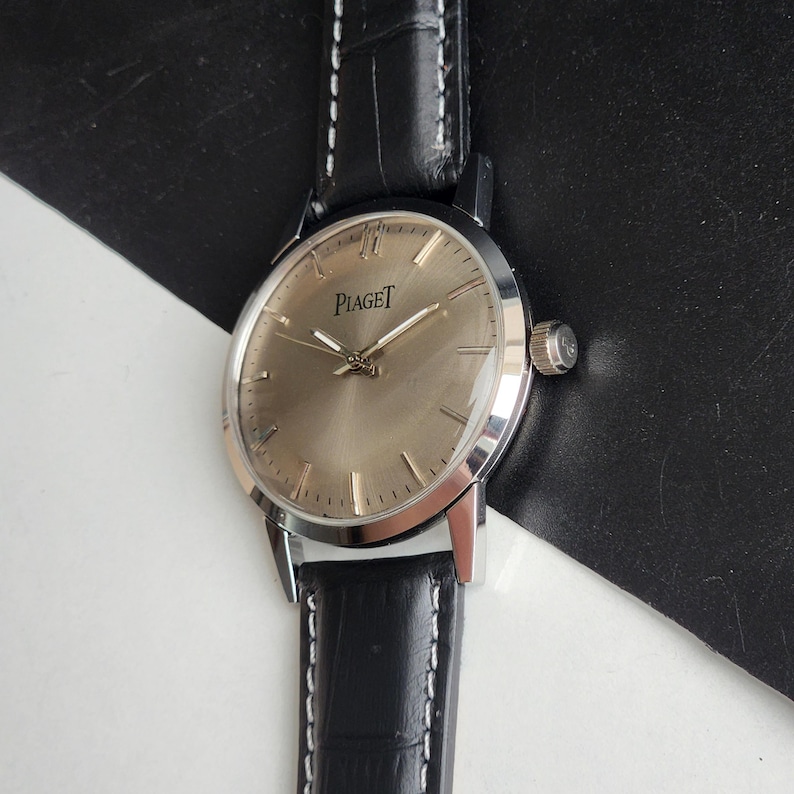 Puede incluir: Un reloj Piaget plateado con esfera marr&oacute;n claro y correa de cuero negro. El reloj tiene una esfera redonda con agujas e indicadores plateados. La correa es de cuero negro con costuras blancas. Dise&ntilde;o cl&aacute;sico.