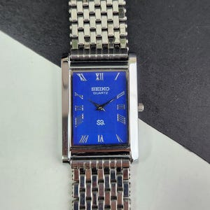 May include: A rectangular Seiko Quartz watch with a blue face and silver-tone metal band. The watch face features Roman numeral hour markers and the Seiko logo. The watch band is a woven style.