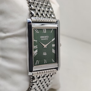 Puede incluir: Un reloj Seiko Quartz rectangular con una correa de metal plateado y una esfera verde. La esfera del reloj muestra números romanos y el logotipo de Seiko Quartz. El reloj se presenta sobre una superficie blanca.