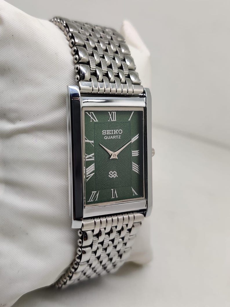 Może przedstawiać: Prostokątny zegarek Seiko Quartz ze srebrną metalową bransoletą i zieloną tarczą. Tarcza zegarka posiada rzymskie cyfry i logo Seiko Quartz. Zegarek jest prezentowany na białej powierzchni.