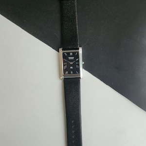 Puede incluir: Un reloj Seiko de cuarzo rectangular con esfera negra y caja plateada. El reloj presenta números romanos y una correa de cuero negro. El reloj se muestra sobre un fondo blanco y negro.