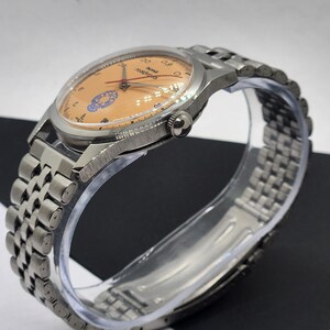 May include: A vintage wristwatch with a silver-toned metal band and case. The watch face is a light orange color with black numerals and hands. The watch has a small crown and a clear crystal. The band is made of small, connected metal links.