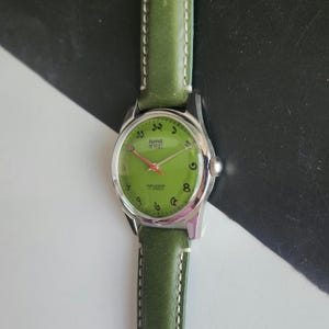 May include: A vintage wristwatch with a green face and a green leather band. The watch has silver casing and Arabic numerals. The watch face has text in a foreign language. The band is stitched with white thread.