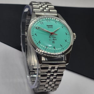 May include: A silver-toned wristwatch with a turquoise face and a matching silver band. The watch face features black numerals and text in a foreign language, with red hands. The watch is a classic design.