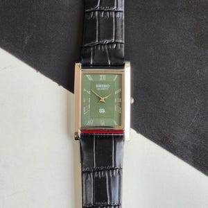 以下が含まれることがあります： 緑色の文字盤と金色のケースを備えた長方形のセイコー クォーツ腕時計。時計はクロコダイル柄の黒革バンドが付いています。文字盤にはローマ数字とセイコー クォーツのロゴが表示されています。