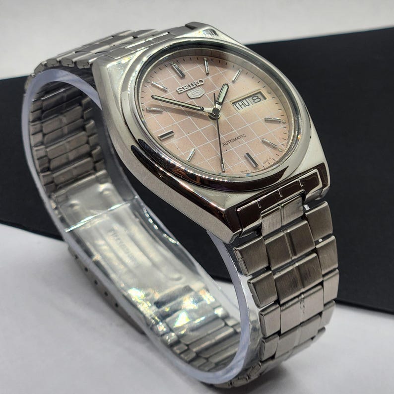 May include: A silver-toned Seiko automatic wristwatch with a pink, grid-patterned face. The watch features a day and date display and a silver metal band. The watch is displayed on a clear plastic stand.