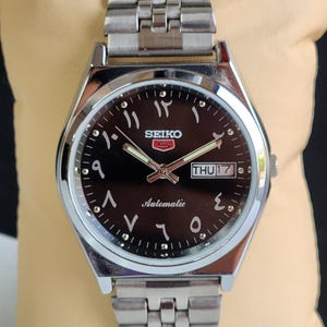 May include: A silver Seiko 5 automatic wristwatch with a black dial and Arabic numerals. The watch has a day and date display and a stainless steel strap. The watch is displayed on a beige surface.