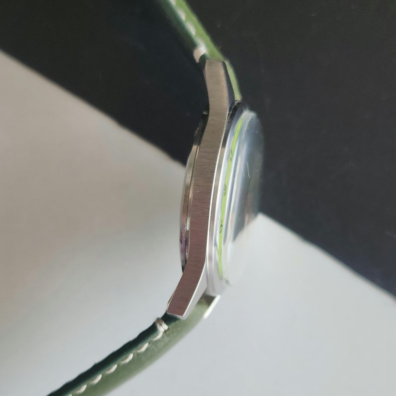May include: A silver-toned wristwatch with a green leather band. The watch face is visible, with a light green ring around the edge. The watch is shown from the side, highlighting its sleek design and the texture of the band.