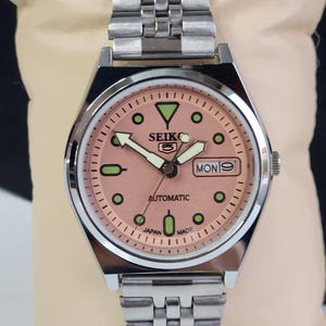 K&ouml;nnte beinhalten: Eine Seiko 5 Automatik-Uhr aus Edelstahl mit einem lachsfarbenen Zifferblatt. Die Uhr hat ein silbernes Armband, gr&uuml;ne und schwarze Stundenmarkierungen und die Tages- und Datumsanzeige auf 3 Uhr. Die Uhr ist mit "Japan Made" gekennzeichnet.