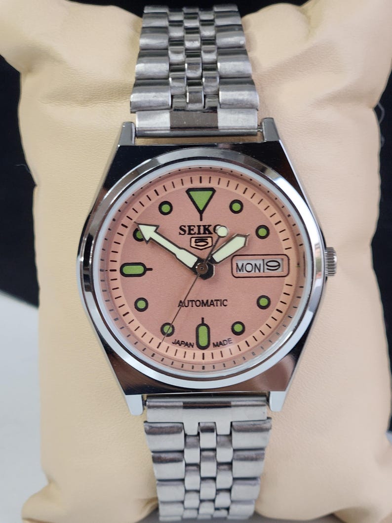 K&ouml;nnte beinhalten: Eine Seiko 5 Automatik-Uhr aus Edelstahl mit einem lachsfarbenen Zifferblatt. Die Uhr hat ein silbernes Armband, gr&uuml;ne und schwarze Stundenmarkierungen und die Tages- und Datumsanzeige auf 3 Uhr. Die Uhr ist mit "Japan Made" gekennzeichnet.
