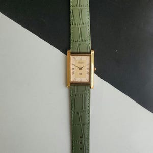 May include: A rectangular gold-toned Seiko watch with a light green leather band. The watch face is cream-colored with gold hands and Roman numeral hour markers. The band has a crocodile skin texture.