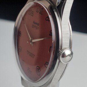 Puede incluir: Primer plano de un reloj de pulsera vintage con esfera granate y caja y correa plateadas. La esfera del reloj presenta n&uacute;meros y agujas negros. El reloj tiene un acabado texturizado y cepillado en la caja.