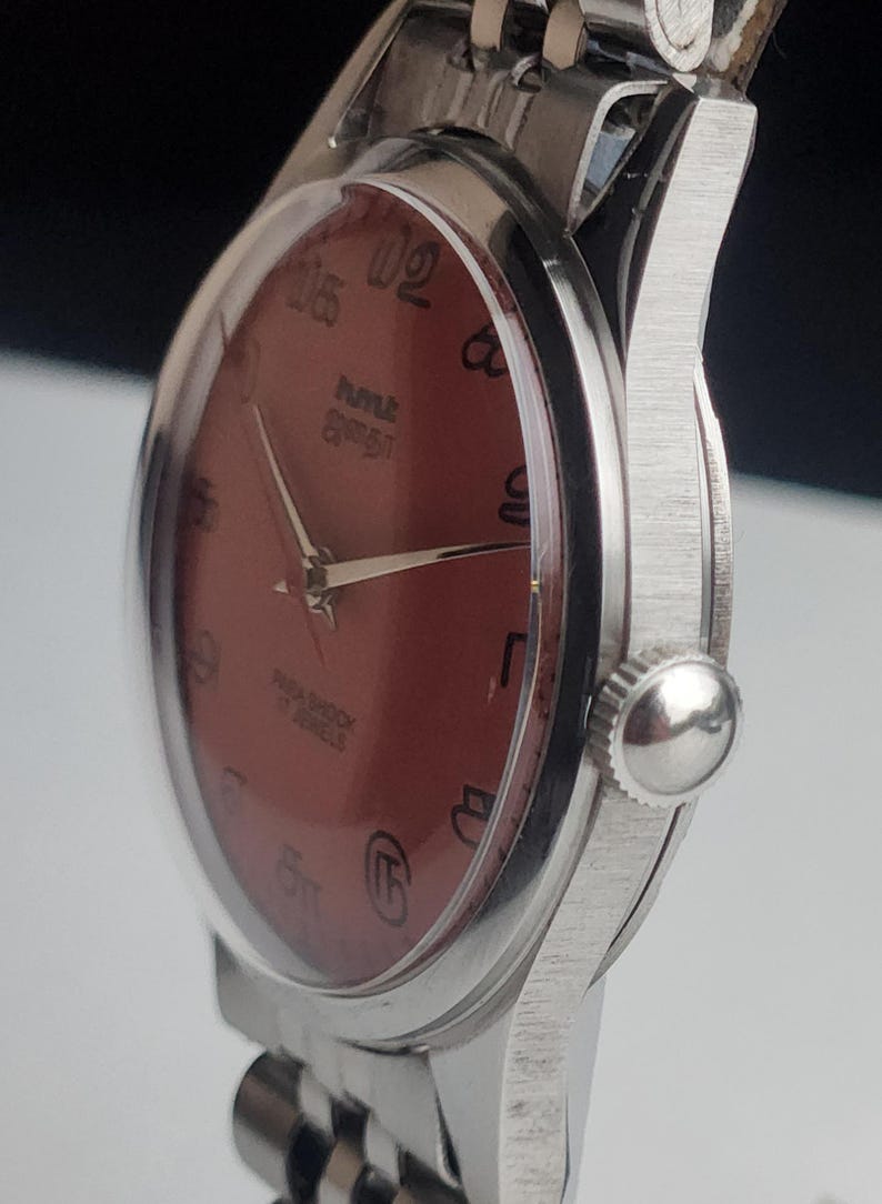 Puede incluir: Primer plano de un reloj de pulsera vintage con esfera granate y caja y correa plateadas. La esfera del reloj presenta n&uacute;meros y agujas negros. El reloj tiene un acabado texturizado y cepillado en la caja.