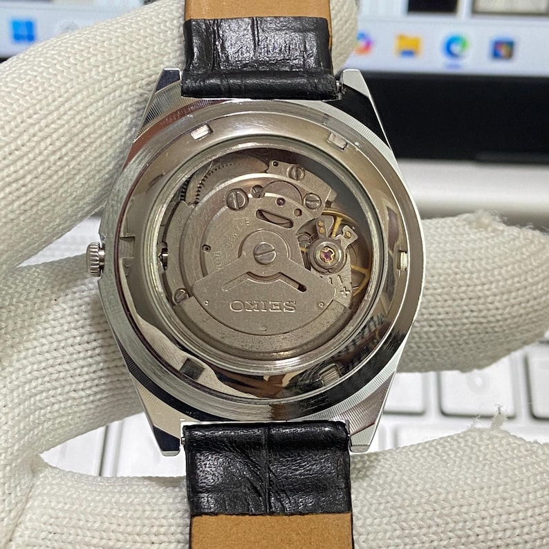 May include: Close-up of a silver-toned watch with a black leather band. The watch's back is transparent, revealing the intricate internal mechanism, including gears and the "SEIKO" brand name. The watch is on a white surface.
