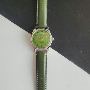 May include: A vintage wristwatch with a green leather band and a silver-colored case. The watch face is green with Arabic numerals and red hands. The watch band is approximately 0.75 inches wide.