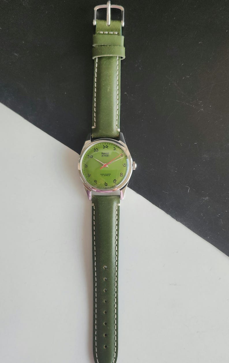 May include: A vintage wristwatch with a green leather band and a silver-colored case. The watch face is green with Arabic numerals and red hands. The watch band is approximately 0.75 inches wide.