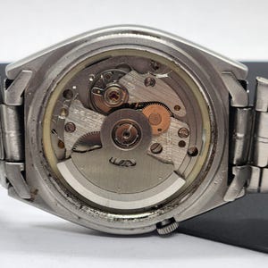 May include: Close-up of the back of a silver-toned wristwatch, showcasing the intricate internal mechanism. The watch's gears, springs, and other components are visible through a clear circular window. The watch band is a silver-toned metal.