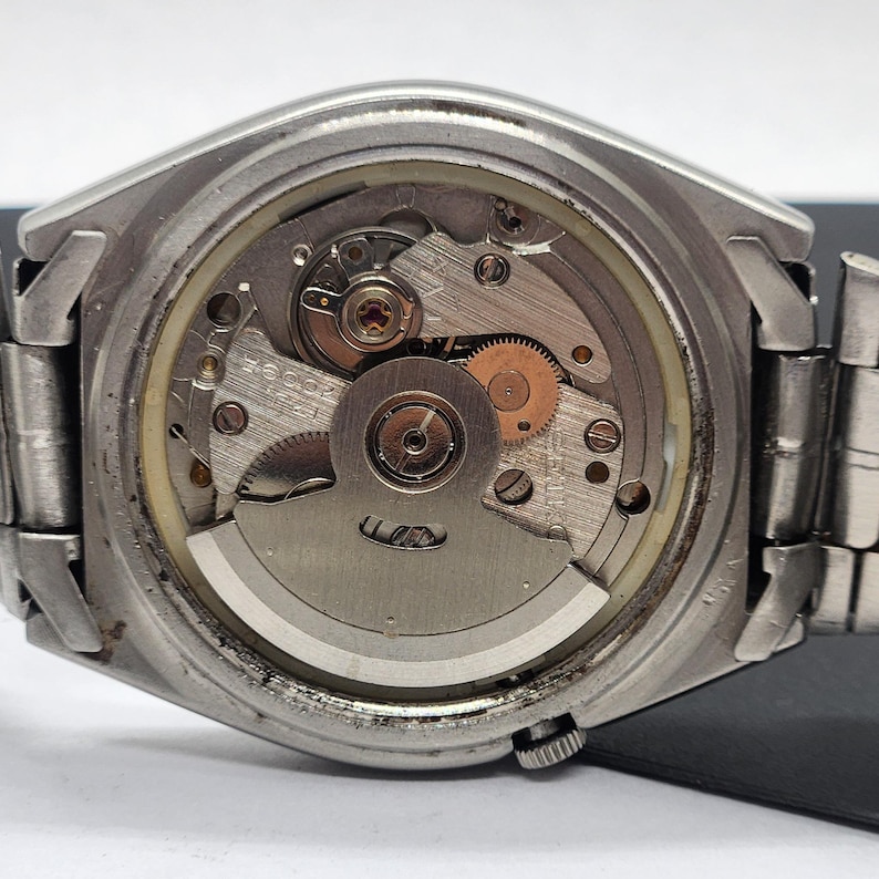 May include: Close-up of the back of a silver-toned wristwatch, showcasing the intricate internal mechanism. The watch's gears, springs, and other components are visible through a clear circular window. The watch band is a silver-toned metal.