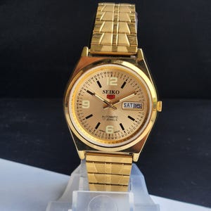 May include: A gold Seiko automatic wristwatch with a gold bracelet strap. The watch face is gold with black numerals and hands, and a day-date display. The watch has a 17 jewels movement and is made in Japan.