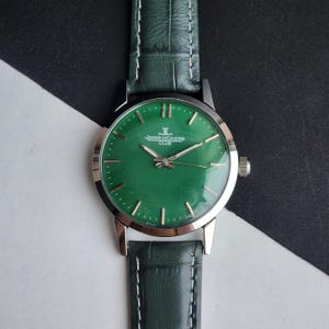 May include: A green-faced Jaeger-LeCoultre Club watch with a silver case and green leather band. The watch face features silver hour markers and hands. The band is green with white stitching.