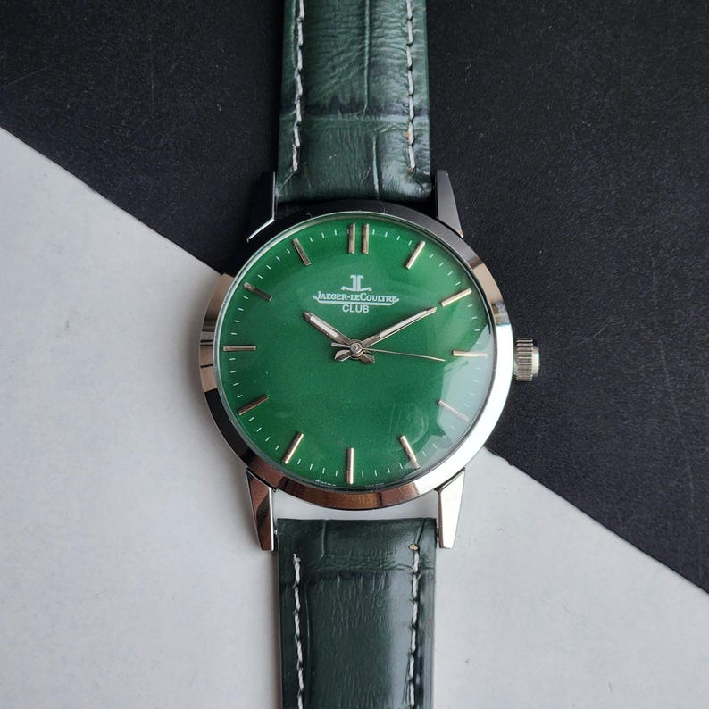 May include: A green-faced Jaeger-LeCoultre Club watch with a silver case and green leather band. The watch face features silver hour markers and hands. The band is green with white stitching.