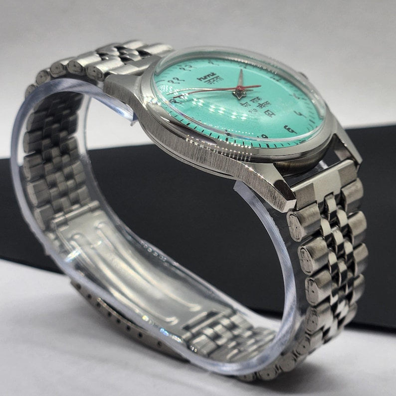 May include: A silver-toned wristwatch with a turquoise face and a matching silver band. The watch face features black numerals and text, with red hands. The band is a linked style, and the watch is displayed on a clear plastic form.
