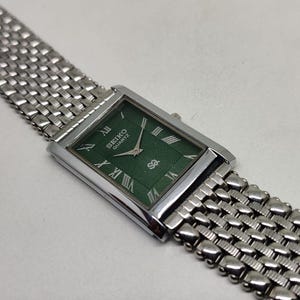 Może przedstawiać: Prostokątny zegarek Seiko Quartz z zieloną tarczą i srebrną metalową bransoletą. Tarcza zegarka posiada rzymskie cyfry i logo Seiko. Bransoleta ma pleciony wz&oacute;r.