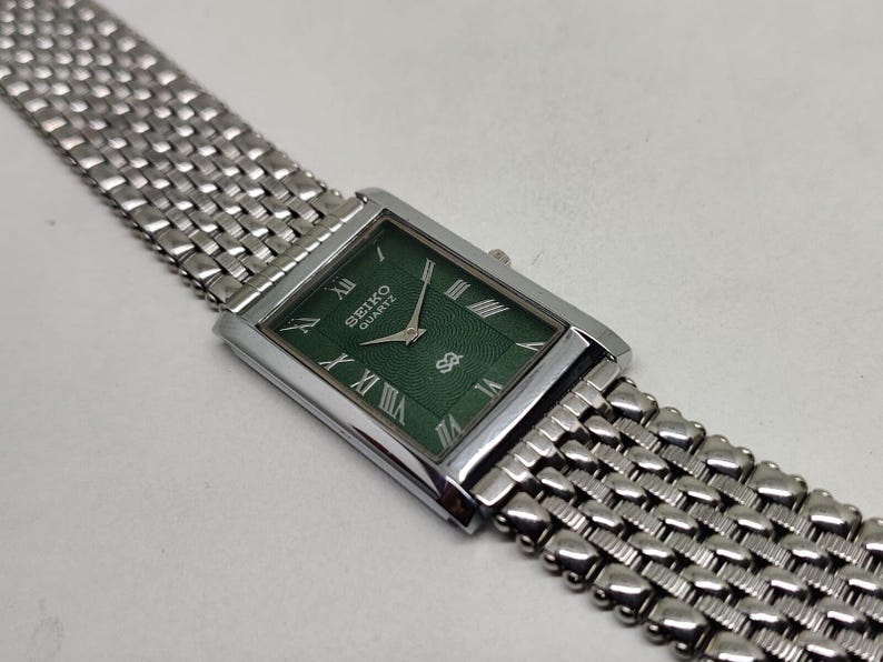 Może przedstawiać: Prostokątny zegarek Seiko Quartz z zieloną tarczą i srebrną metalową bransoletą. Tarcza zegarka posiada rzymskie cyfry i logo Seiko. Bransoleta ma pleciony wz&oacute;r.