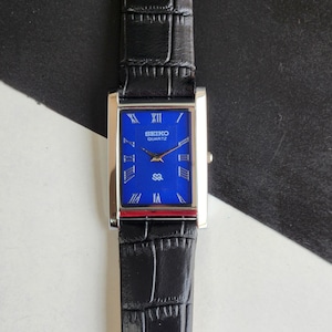 Może przedstawiać: Prostokątny zegarek Seiko Quartz z niebieską tarczą i srebrną kopertą. Zegarek posiada rzymskie cyfry i czarny skórzany pasek. Tarcza zegarka wyświetla logo Seiko Quartz.