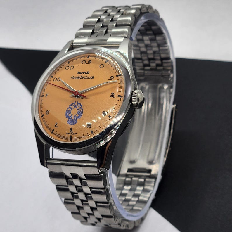 May include: A silver wristwatch with a light orange face and a silver band. The watch face has black numerals, a blue emblem, and red hands. The text "HMT" and "rookiearabool" are printed on the watch face.