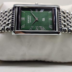 Może przedstawiać: Prostokątny zegarek Seiko Quartz z zieloną tarczą i srebrnymi rzymskimi cyframi. Zegarek ma srebrną obudowę i plecioną metalową bransoletę. Zegarek jest prezentowany na białej powierzchni.