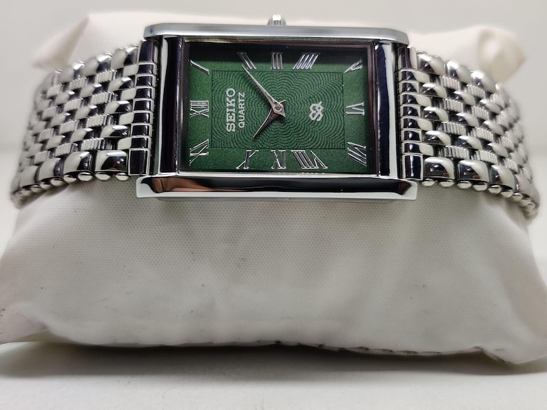 Może przedstawiać: Prostokątny zegarek Seiko Quartz z zieloną tarczą i srebrnymi rzymskimi cyframi. Zegarek ma srebrną obudowę i plecioną metalową bransoletę. Zegarek jest prezentowany na białej powierzchni.