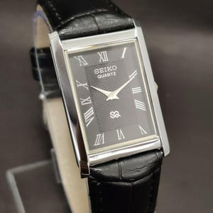 May include: A rectangular Seiko Quartz watch with a black dial and silver case. The watch has Roman numeral hour markers and a black leather strap. The watch face displays the Seiko Quartz logo and a textured central pattern.