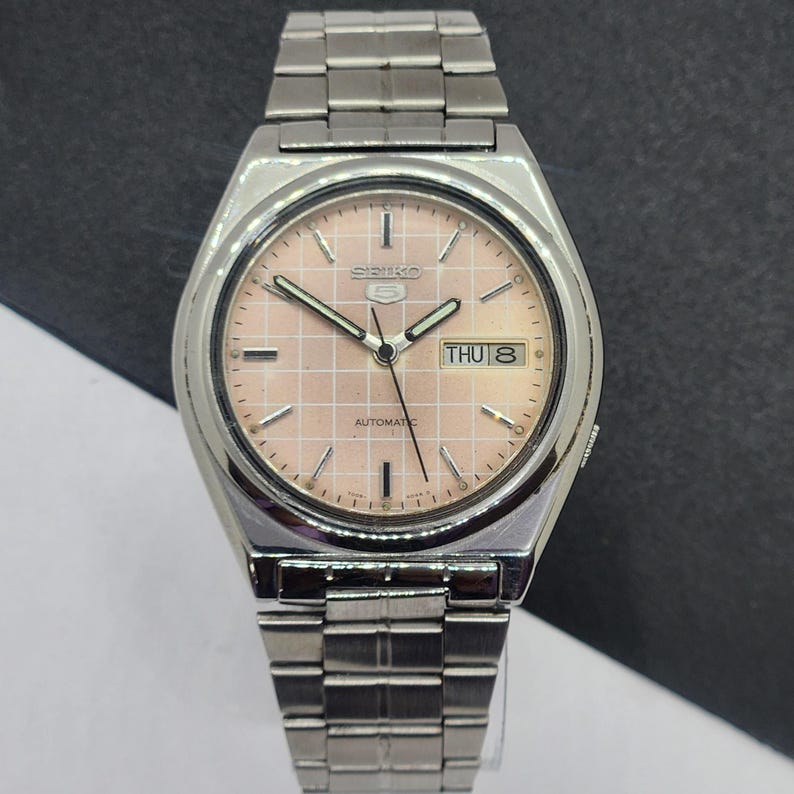 May include: A vintage Seiko 5 automatic wristwatch with a silver-tone stainless steel band and case. The watch features a light pink dial with a grid pattern, day and date display, and silver hands. The watch face reads "Seiko 5 Automatic".