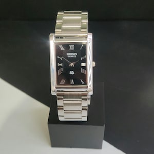 May include: A rectangular Seiko Quartz watch with a silver metal strap and a black face. The watch face has Roman numerals and the Seiko Quartz logo. The watch is displayed on a black stand.