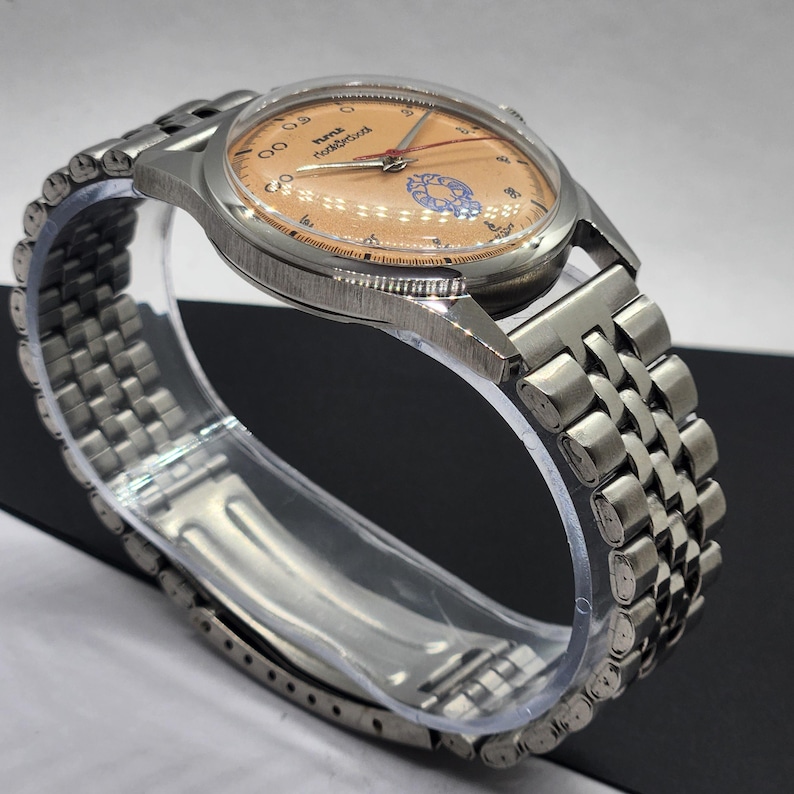 May include: A stainless steel wristwatch with a peach-colored face and a silver-tone metal band. The watch face features black numerals and hands, with the brand name "Hymex" and the words "Nodelbrot" printed on the face. The watch is displayed on a clear plastic stand.