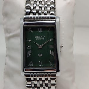 Może przedstawiać: Prostokątny zegarek Seiko Quartz ze srebrną kopertą i bransoletą. Tarcza zegarka jest ciemnozielona ze srebrnymi rzymskimi cyframi i wskaz&oacute;wkami. Bransoleta składa się z małych, połączonych metalowych ogniw.